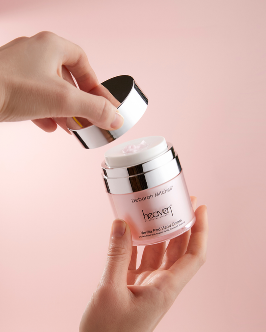Hand holding a pink jar of Deborah Mitchell Heaven Vanilla Pod Hand Cream against a light pink background