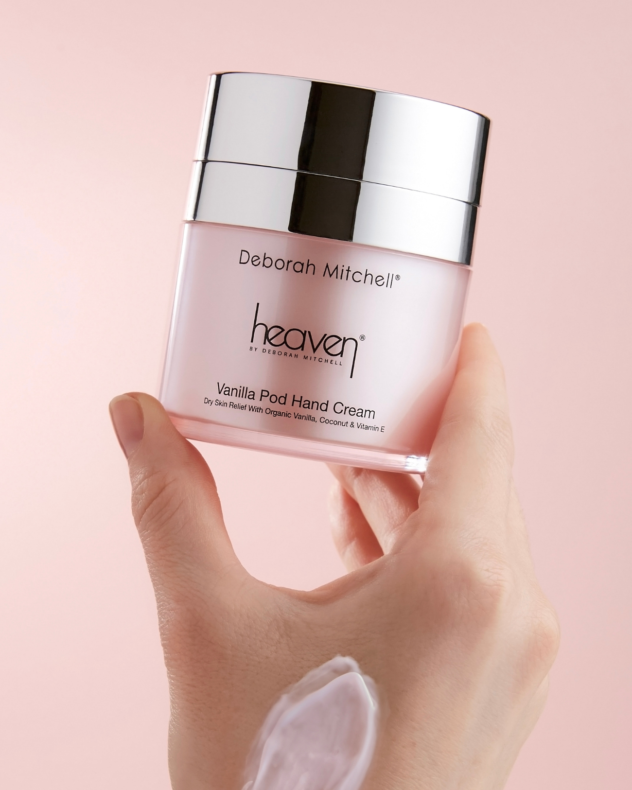 Hand holding a pink jar of Deborah Mitchell heaven Vanilla Ped Hand Cream against a pink background