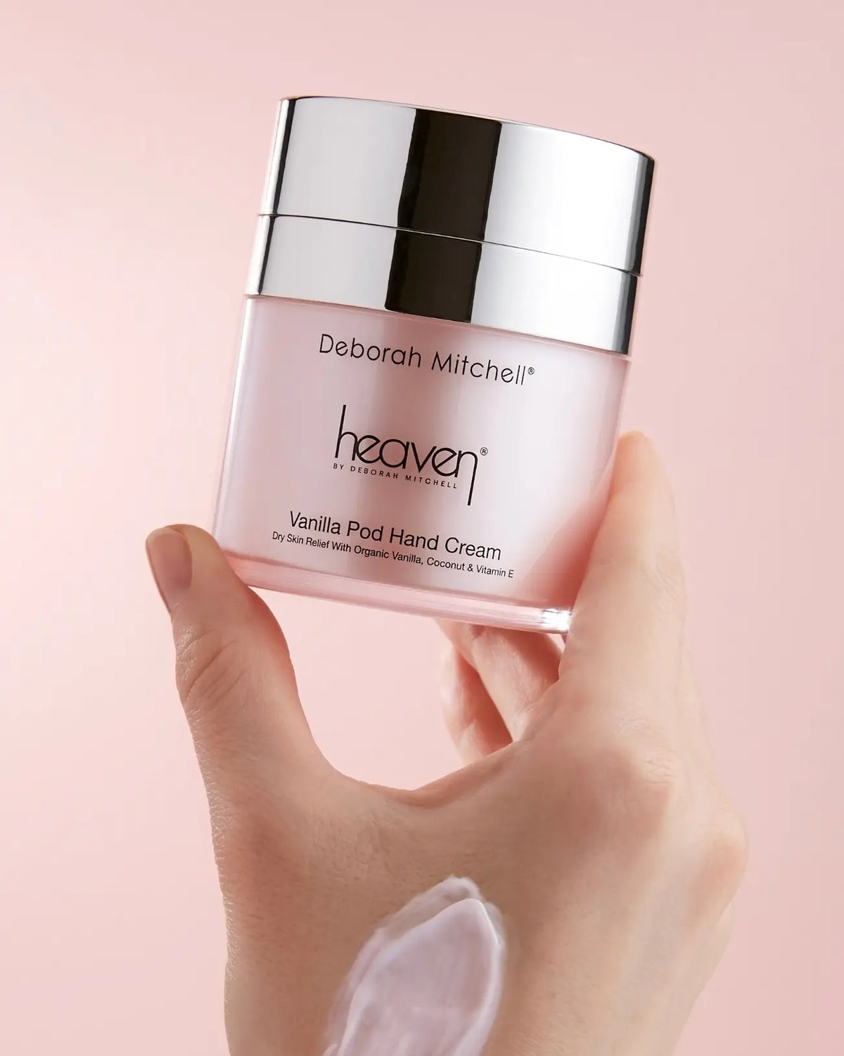 Hand holding a pink jar of Deborah Mitchell heaven Vanilla Ped Hand Cream against a pink background