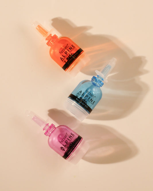 Three dropper bottles with colorful caps on a beige background. Bottles appear in pink, blue and orange.
