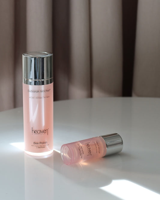 Two pink skincare bottles with silver caps on a white surface. Beige colored curtain in the background.
