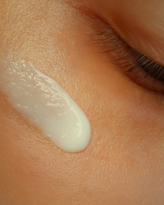 A close-up of a creamy white Formula Bee Eyes eye cream applied to skin below an eye with long lashes.
